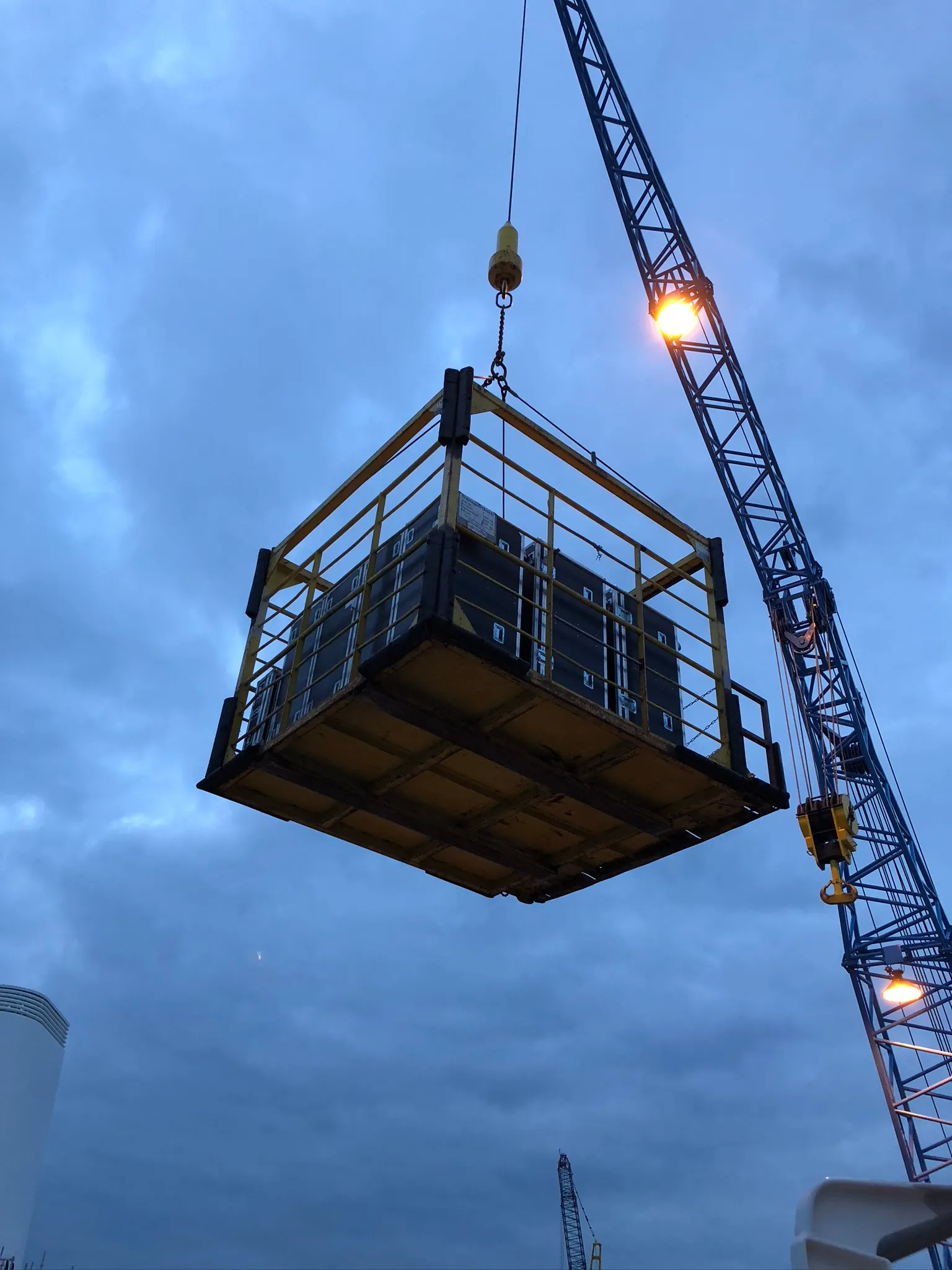 Eine Materialkiste hängt am Kranhaken vor dem Abendhimmel über dem Oberdeck